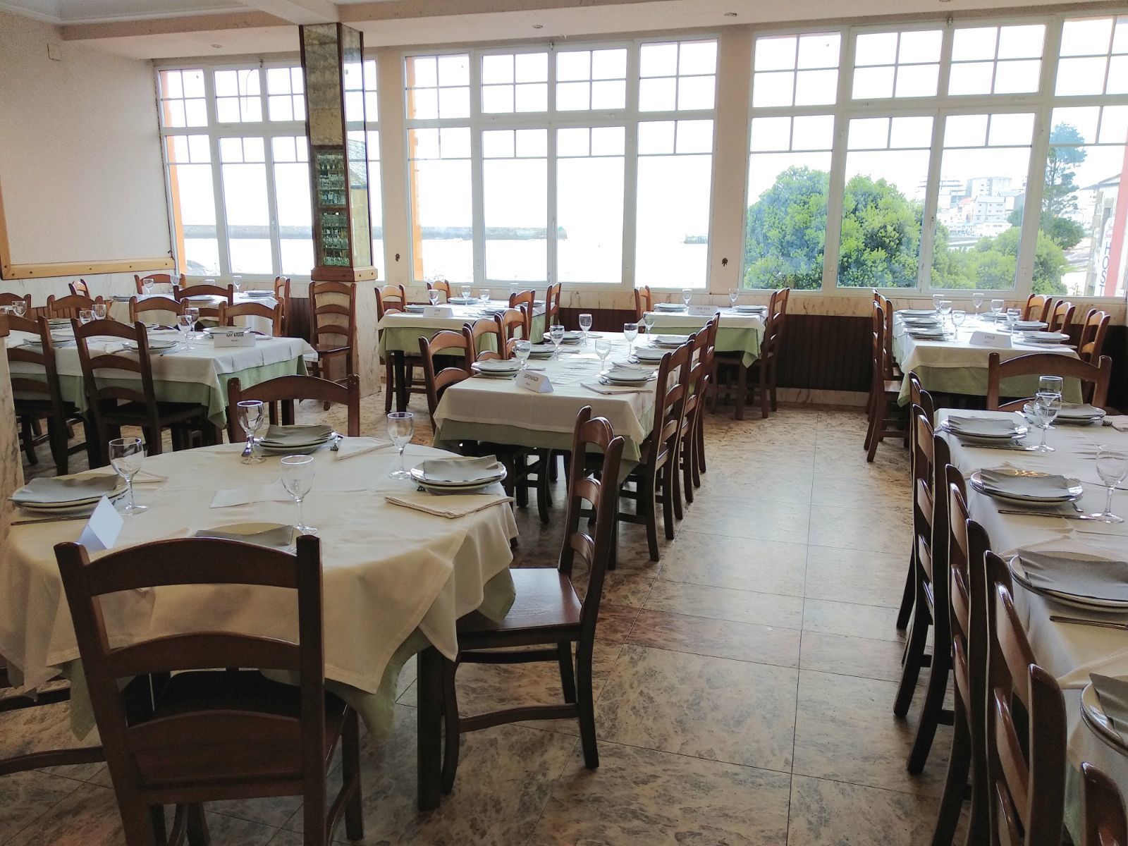 Un restaurant avec de nombreuses tables dressées pour le service, de grandes fenêtres et une vue sur l’extérieur.