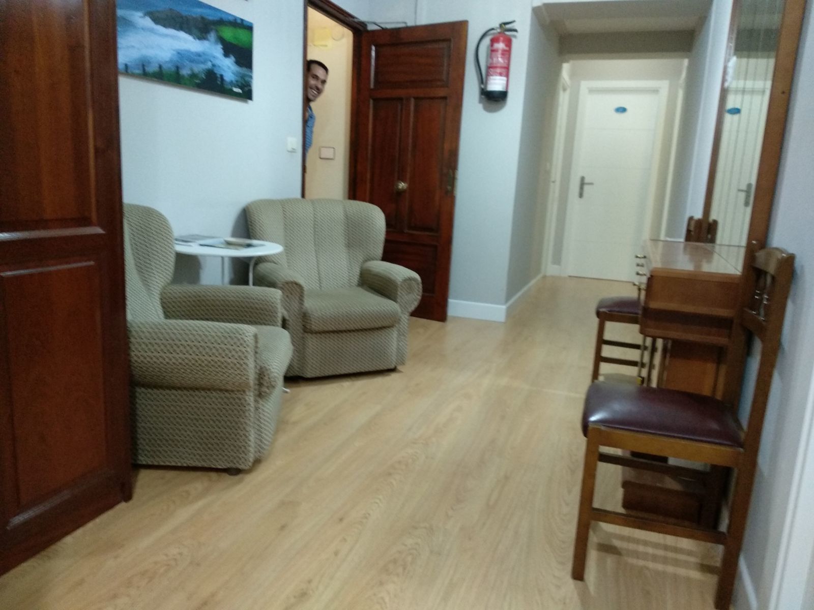 A room with a wooden floor, two armchairs, a small round table, a wooden chair, a door, and a fire extinguisher on the wall.