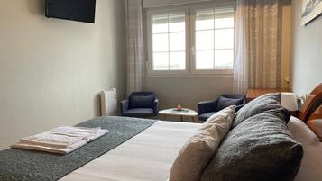 A hotel room with a bed, two armchairs, a small table, a TV mounted on the wall, and a window with curtains.