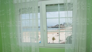 A room with a large window offering a view of a beach and buildings across the water. - Hostal Buenavista (Cervo)