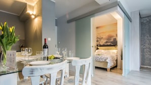 A dining area with a glass table, white chairs, and a wine bottle. There is a bed with a patterned bedspread in the background. - Apartamenty hoteLOVE przy Strumieniu (Karpacz)