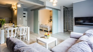 A modern living room with a sofa, a dining table with chairs, a kitchen area with a dining set, and a bedroom with a bed and a TV. - Apartamenty hoteLOVE przy Strumieniu (Karpacz)