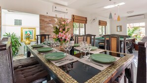 A dining area with a long table set for six, surrounded by cushioned chairs, a decorative vase with flowers, and a pool table in the background. - Madee Villa - Pattaya Holiday House Walking Street (Pattaya)