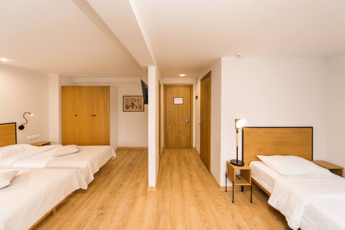 A hotel room with two beds, wooden flooring, a wooden door, a nightstand with a lamp, and a framed picture on the wall.