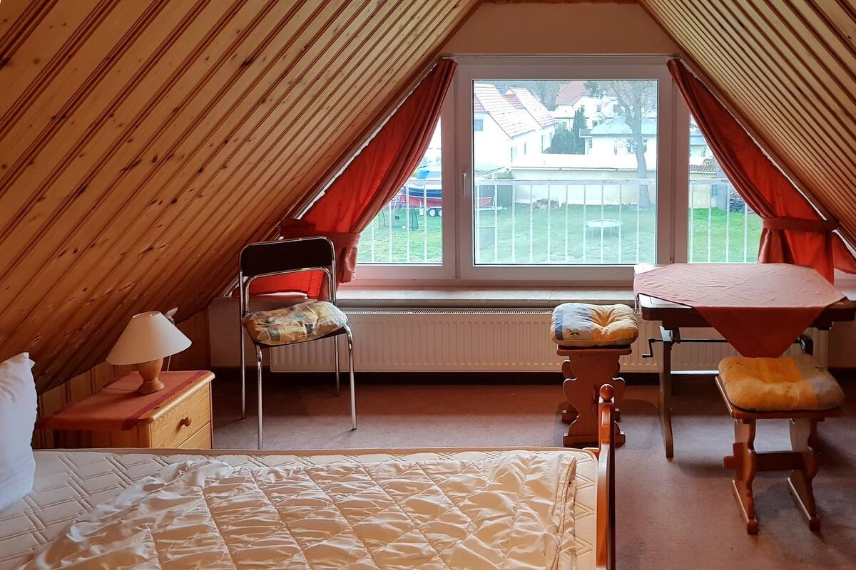 Un dormitori d'àtic acollidor amb un llit, una cadira, una taula i una finestra que donava a una zona residencial.