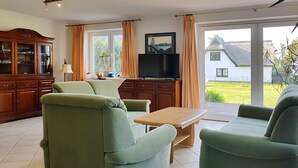 A living room with green sofas, a wooden cabinet, a television, and a view of a house and garden through large windows. - Ferienwohnungen An der Sonnenwiese (Ostseebad Prerow)