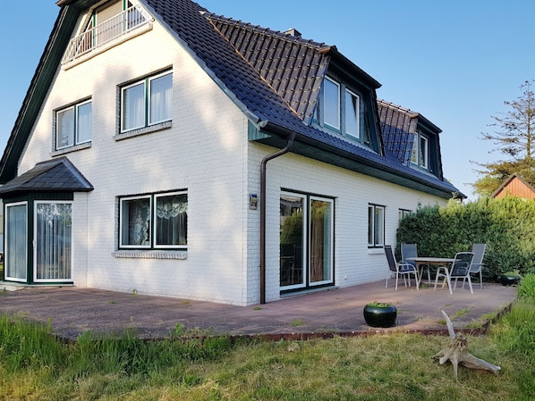 Une maison à deux étages, dotée d’un toit en pente, de grandes fenêtres et d’un patio aménagé avec du mobilier d’extérieur.