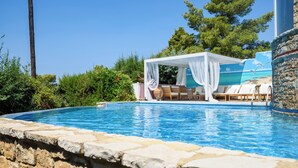 Un espace aménagé au bord de la piscine, comprenant un coin salon couvert, des chaises longues et un mur en pierre.