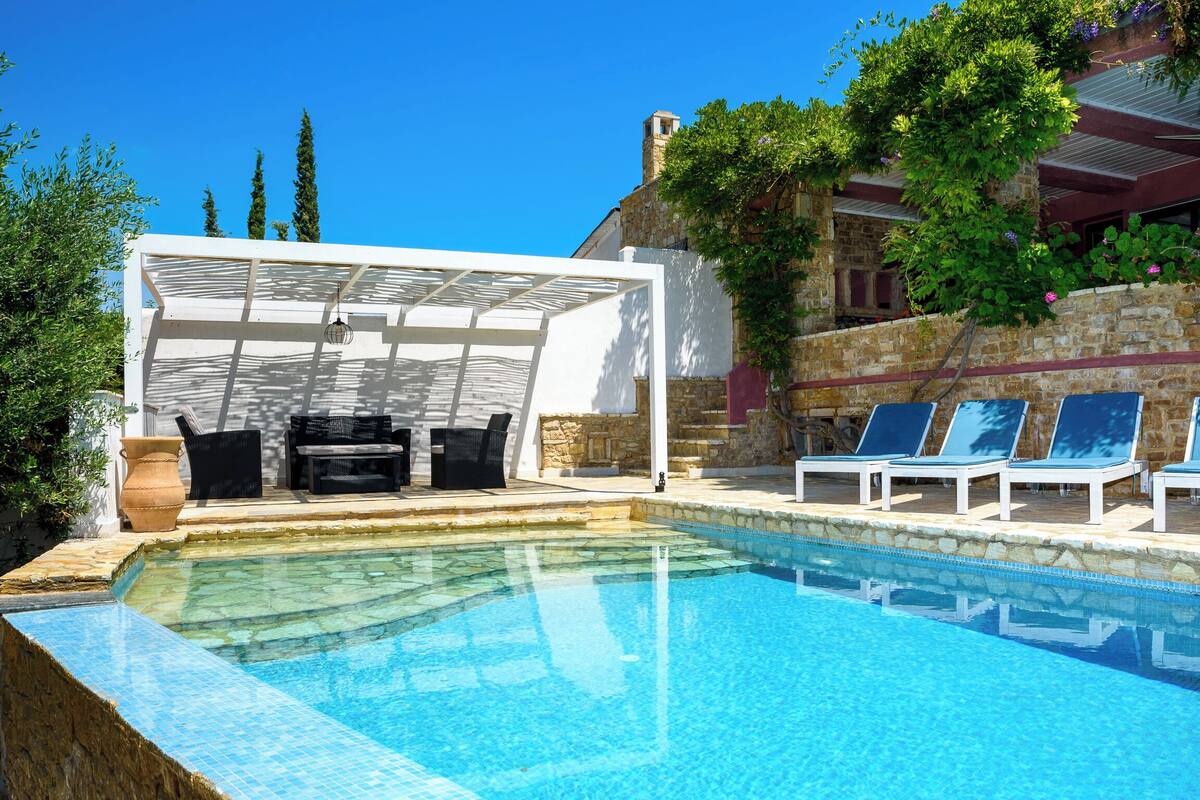 A pool with clear blue water, surrounded by lounge chairs and a covered seating area with outdoor furniture.