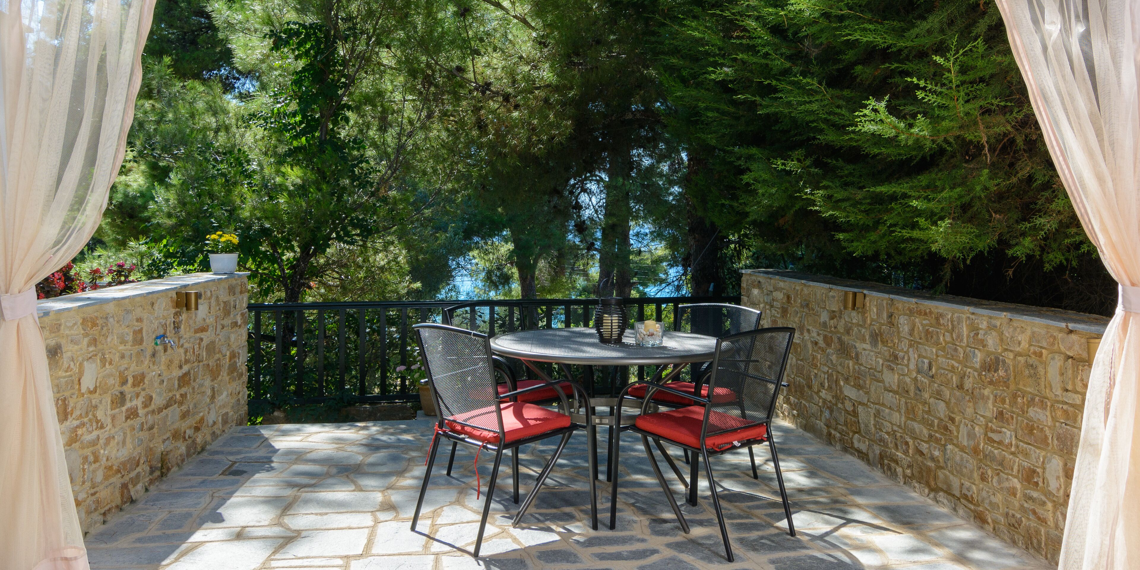 Un patio al aire libre con suelo de piedra, sillas de metal con cojines rojos y una pared de piedra.