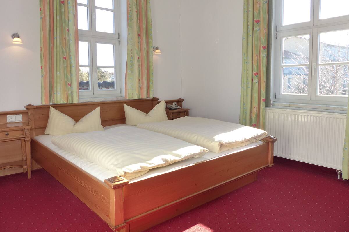 Ein Doppelbett mit Holzrahmen und ein Teppichboden in Rot. Zwei Nachttische mit Lampen sowie ein Fenster mit Vorhängen.
