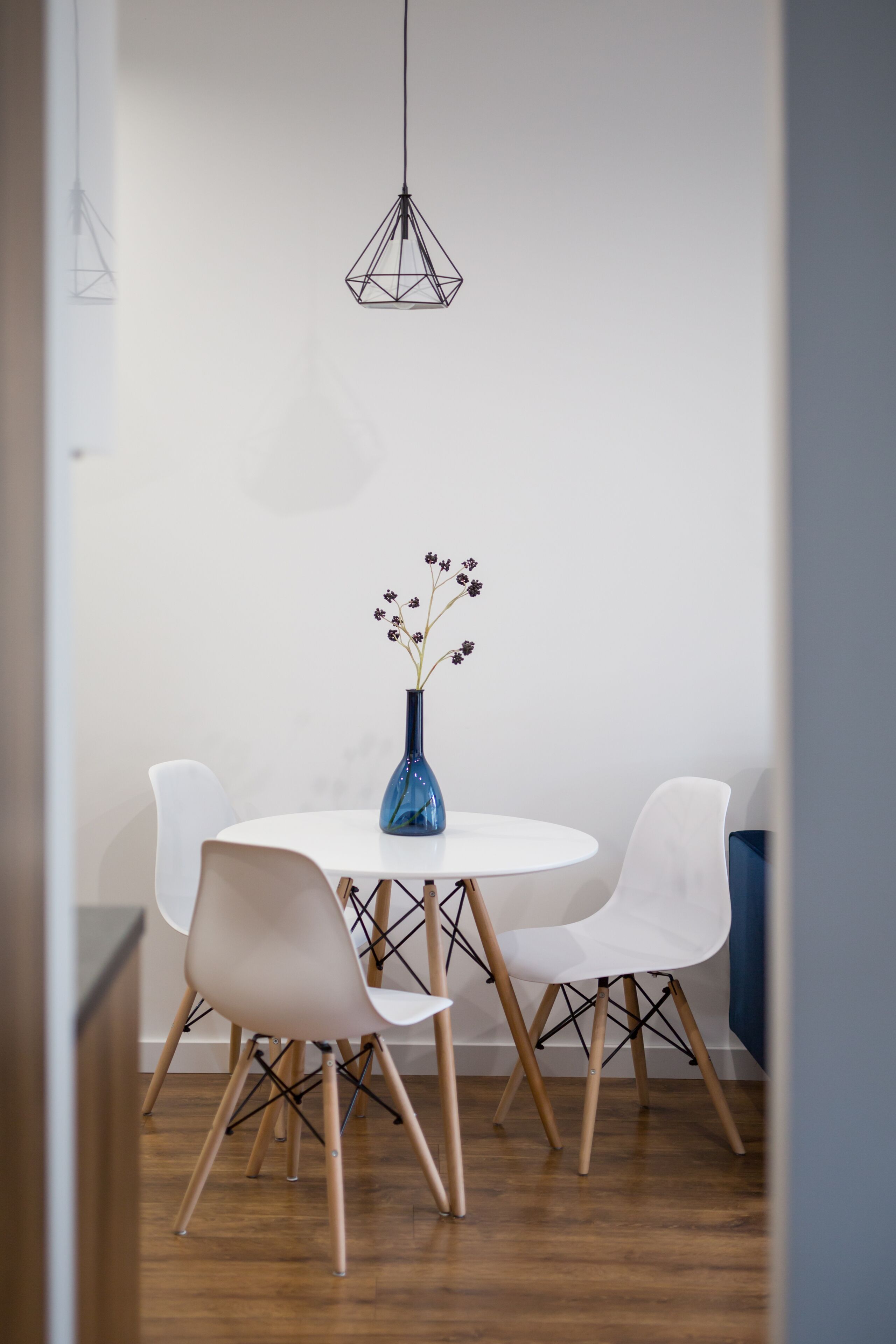 A dining area with a round table, two white chairs, a blue vase with flowers, and a hanging light fixture.