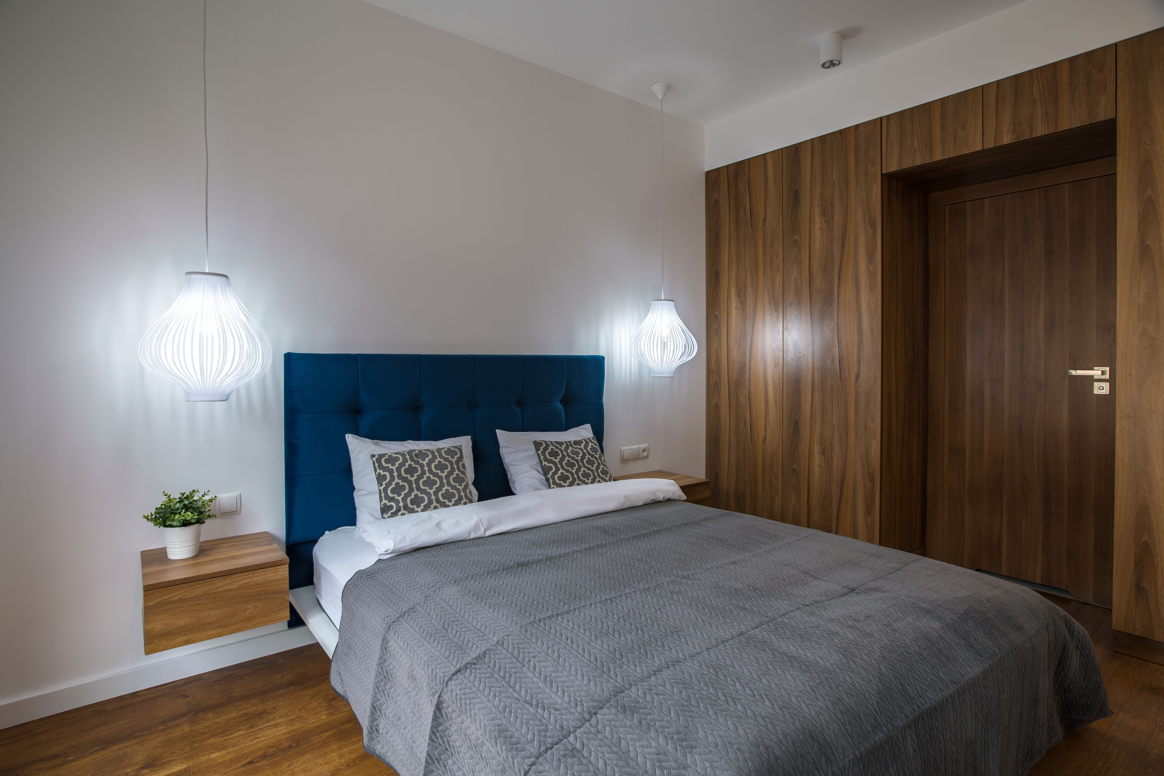 A bedroom with a blue headboard, a grey bedspread, two bedside lamps, a small plant, and a wooden wardrobe.