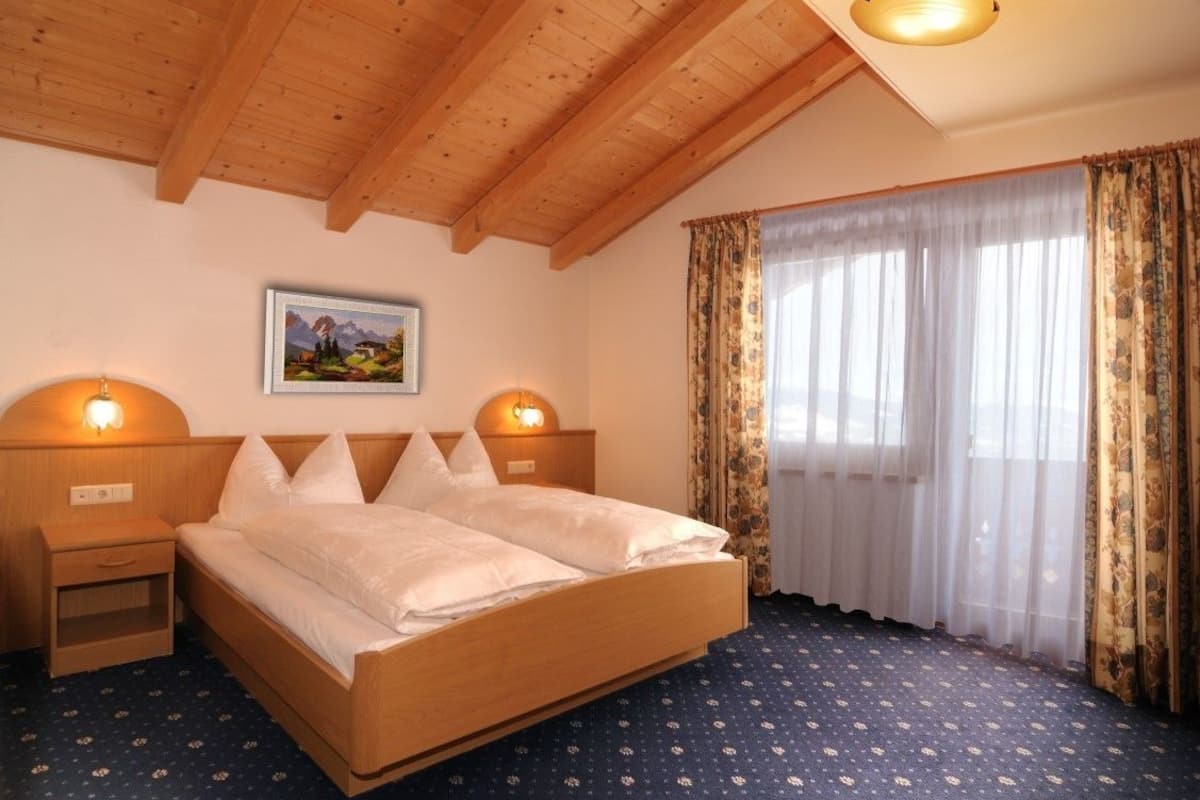Une chambre d’hôtel avec un plafond en bois, un lit recouvert de draps blancs, deux lampes de chevet, une grande fenêtre avec des rideaux légers et un tableau encadré accroché au mur.