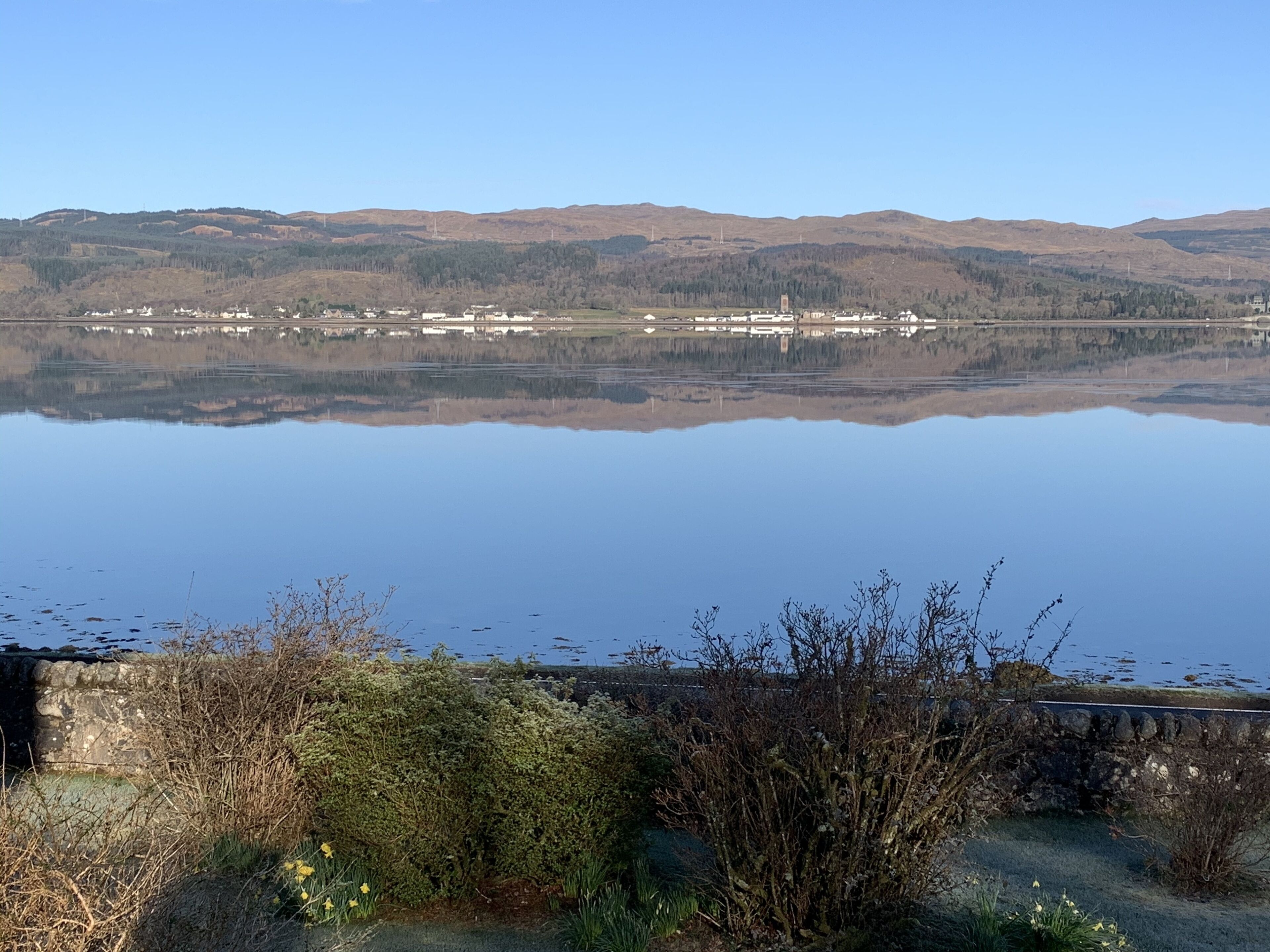 Un lago tranquilo con un reflejo claro del paisaje circundante, que incluye edificios lejanos y colinas.