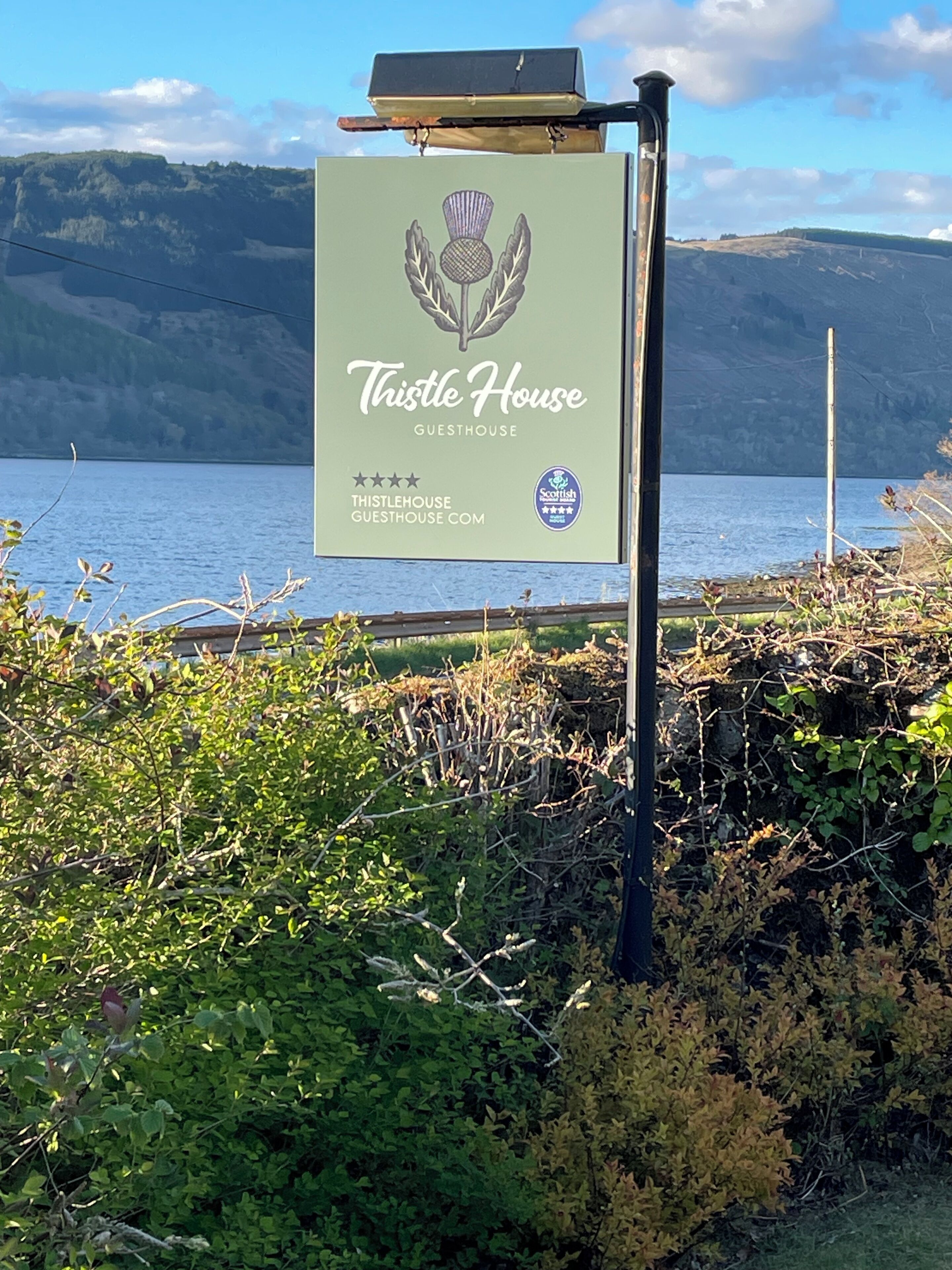 Un letrero para la Casa de Huéspedes Thistle House, con una vista panorámica de un lago al fondo.