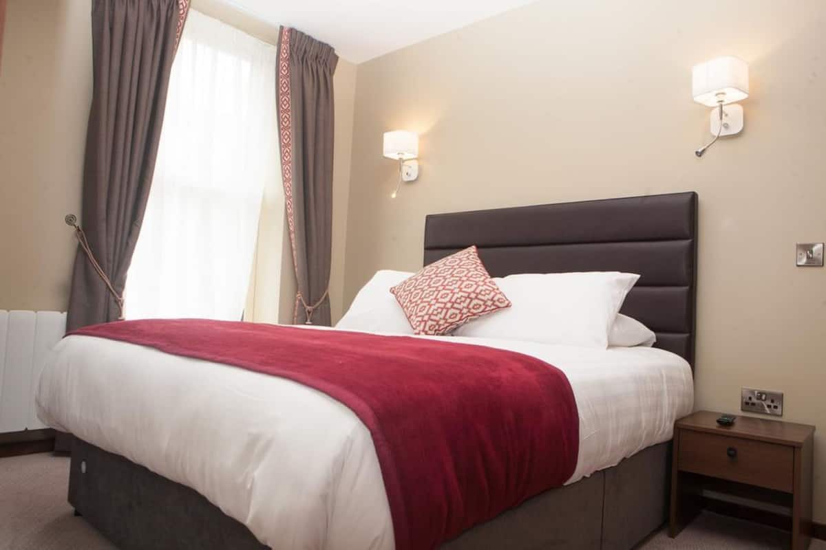 Un lit bien fait, recouvert d’une couverture rouge, avec deux oreillers et une table de chevet sur laquelle se trouve une lampe.