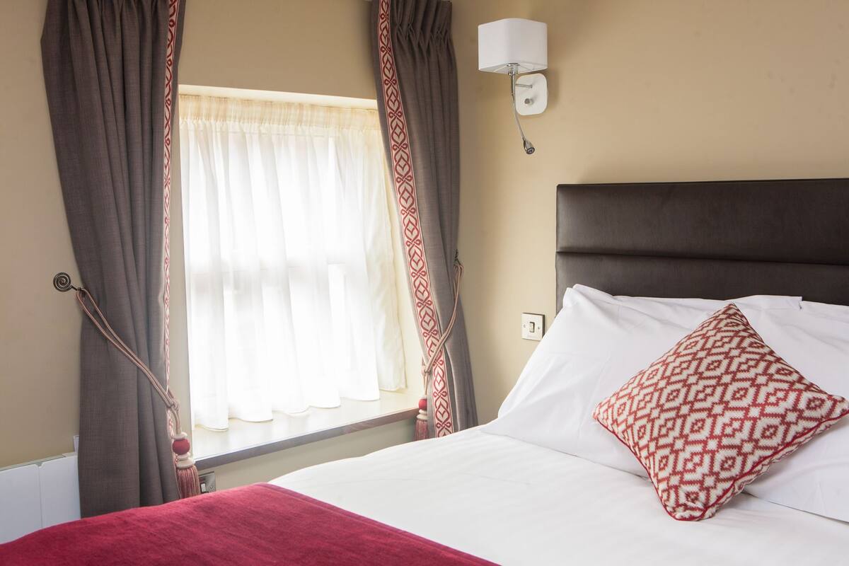 Une chambre d’hôtel avec un lit, une fenêtre avec des rideaux, une lampe fixée au mur et un coussin à motifs.
