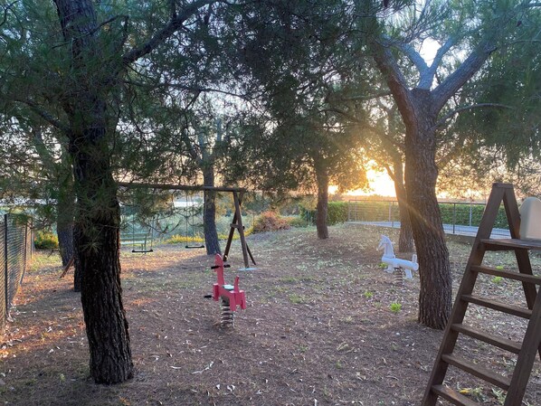 Un terrain de jeux avec des balançoires et un toboggan, entouré d’arbres et d’une clôture.