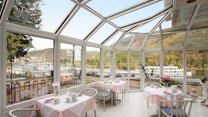 A conservatory with tables set for dining, overlooking a river and hills. - Haus Erholung (Cochem)