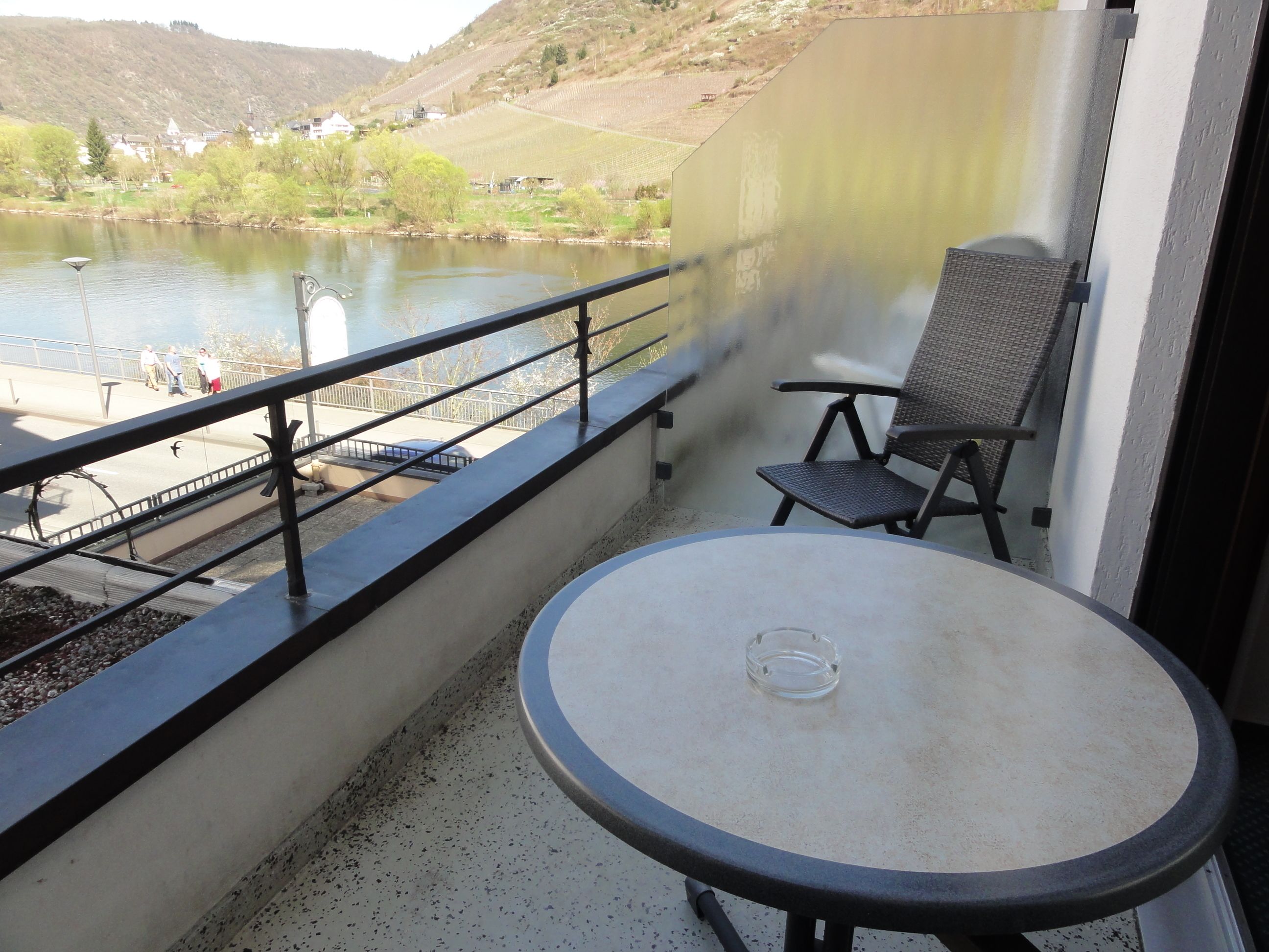 A balcony with a table and chair, overlooking a river and vineyard.