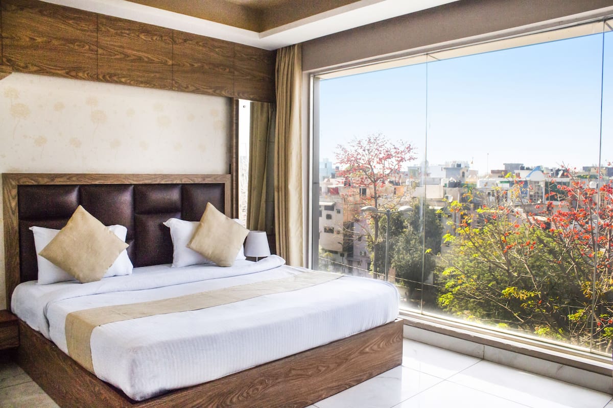 A hotel room with a large bed, wooden headboard, and a view of the city through a large window.