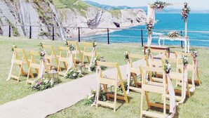 A coastal wedding setup with wooden chairs and a floral arch, overlooking the sea.