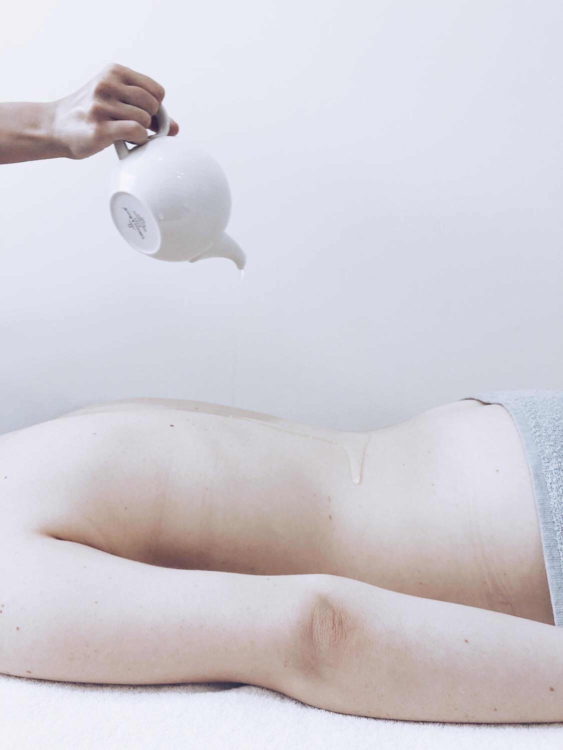 A person lying on a bed with a towel, and a hand pouring liquid from a white container.