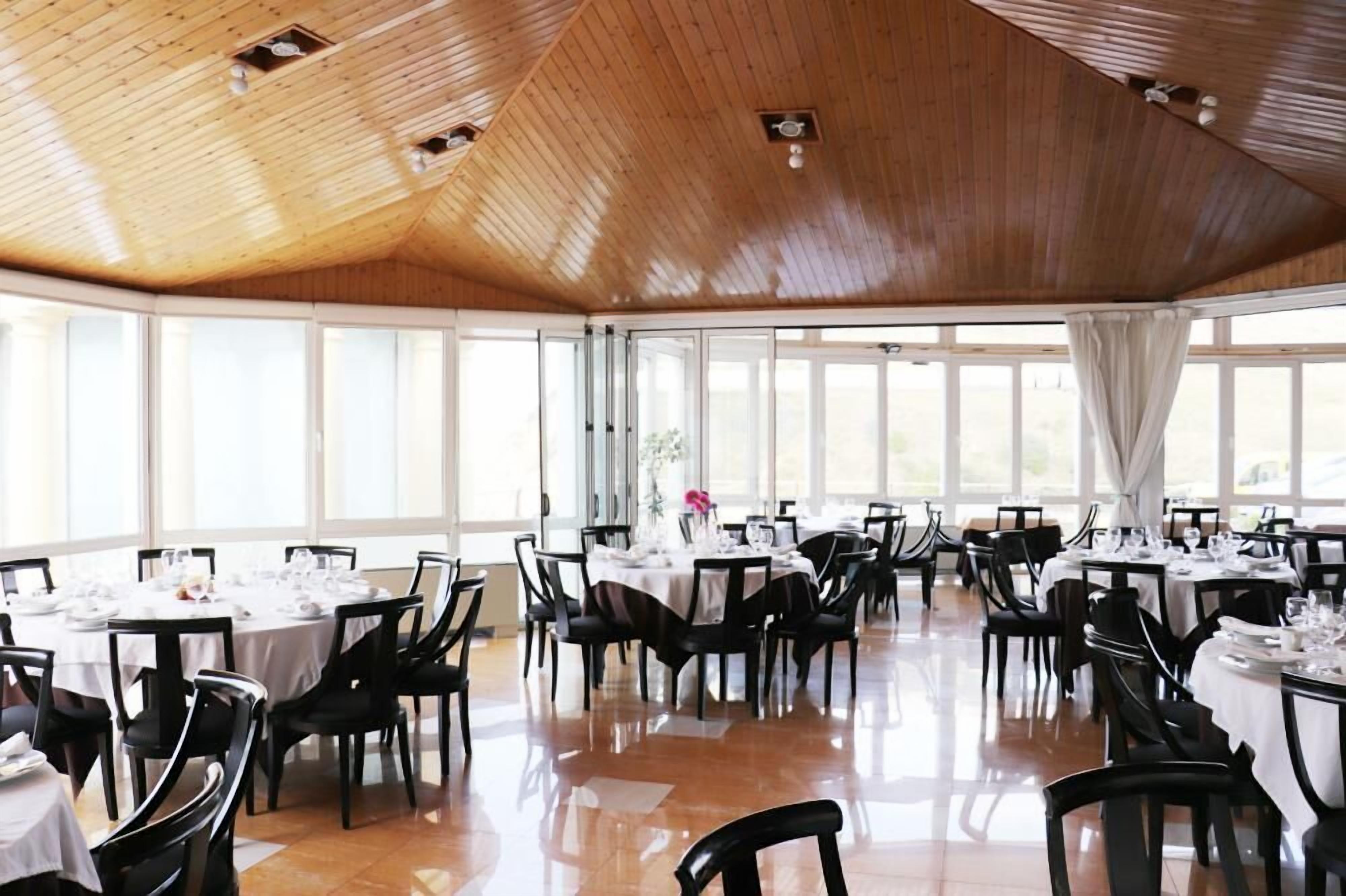 A spacious dining area with round tables and black chairs, large windows, and a wooden ceiling.