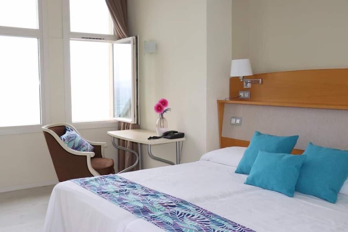 Une chambre d’hôtel comprenant un lit, un bureau, une chaise, une fenêtre et une table de chevet avec une lampe.