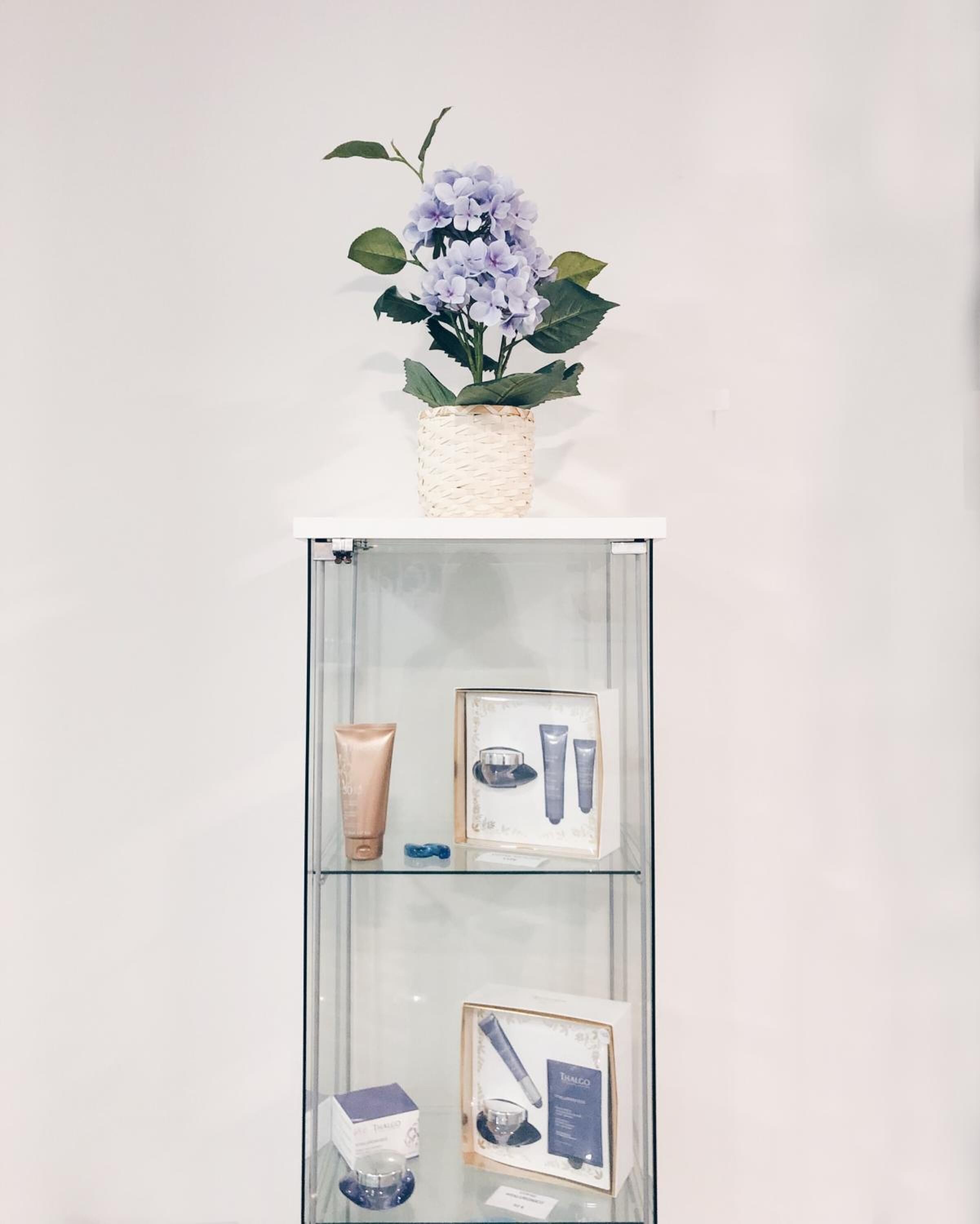 Een displayplank met een gevlochten mand waarin een bloemstuk in paarse tinten staat, en drie ingelijste dozen met huidverzorgingsproducten.