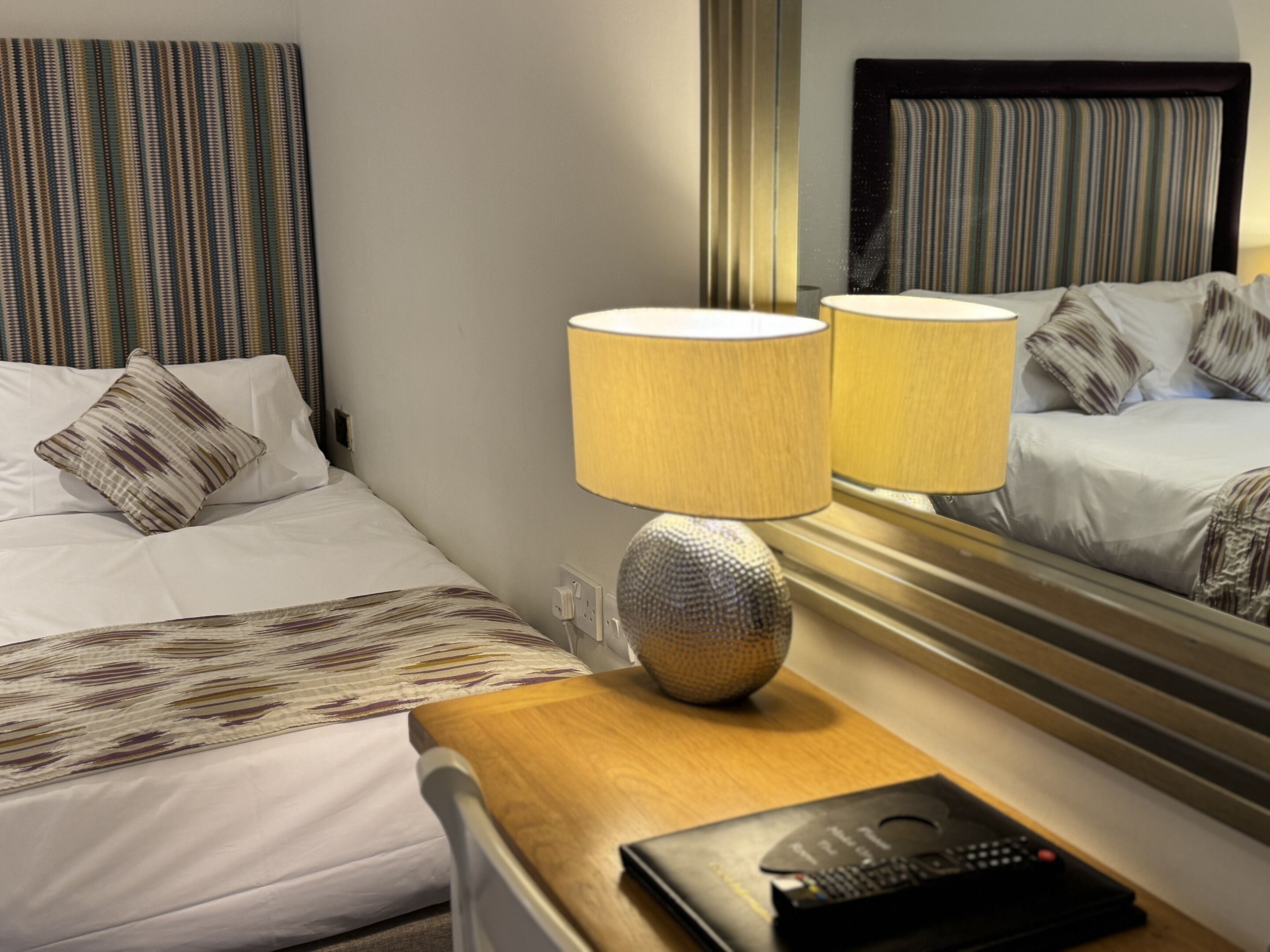 Hotelkamer met een bed, twee bedlampen, een nachtkastje met een afstandsbediening en een spiegel die het bed en de gordijnen weerspiegelt.