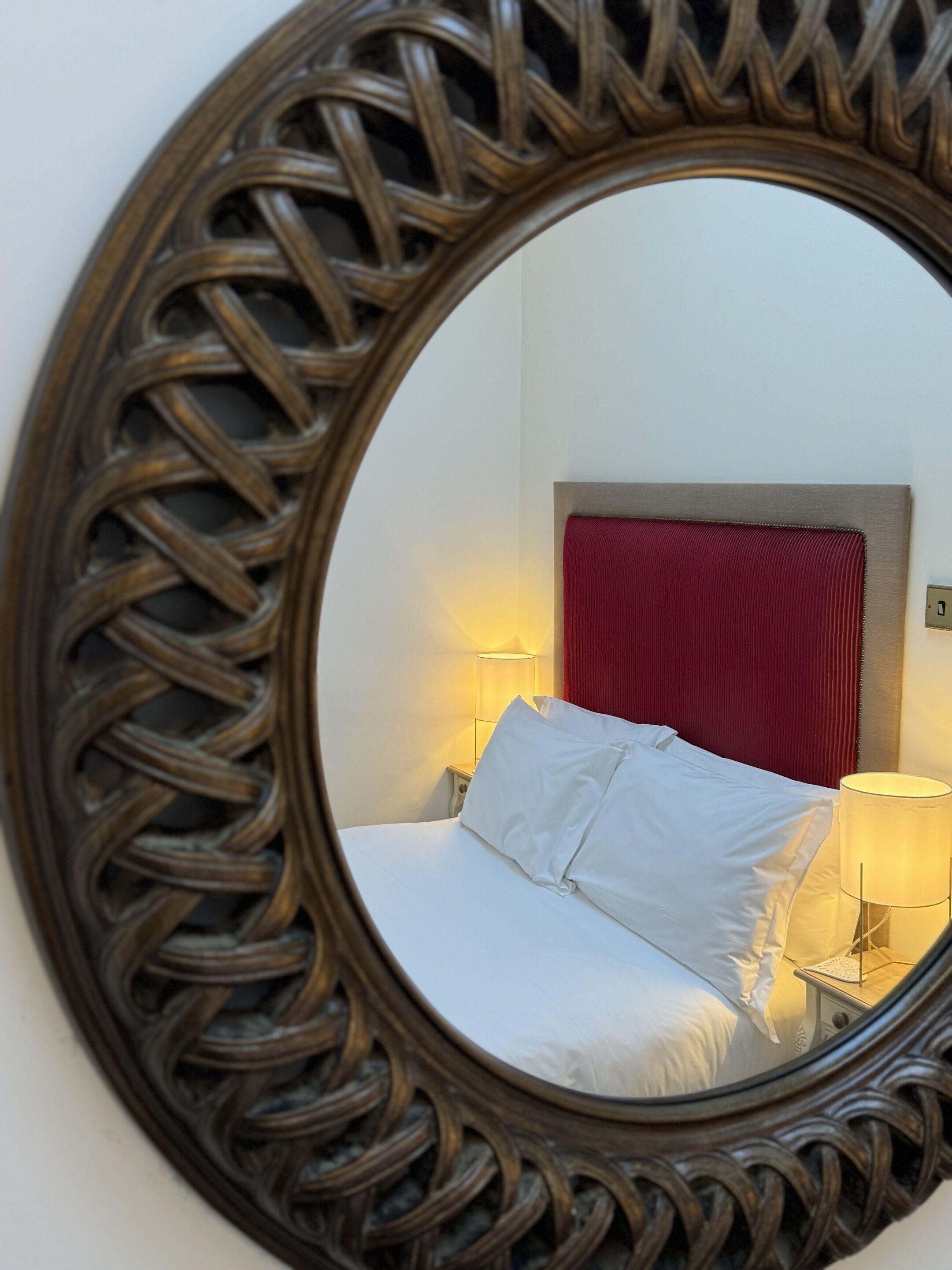 Um espelho com moldura decorativa reflete uma cama, uma cabeceira vermelha e abajures.