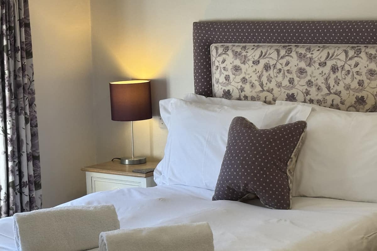 A neatly made bed with white linens and two folded towels, a bedside lamp, and a floral-patterned headboard.