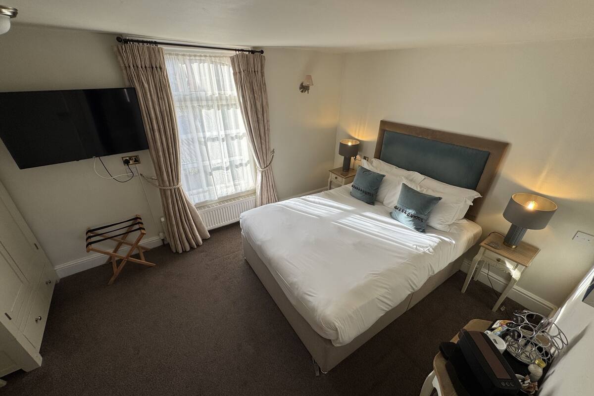 A hotel room with a bed, a flat-screen TV mounted on the wall, a window with curtains, bedside lamps, and a small desk with a lamp.