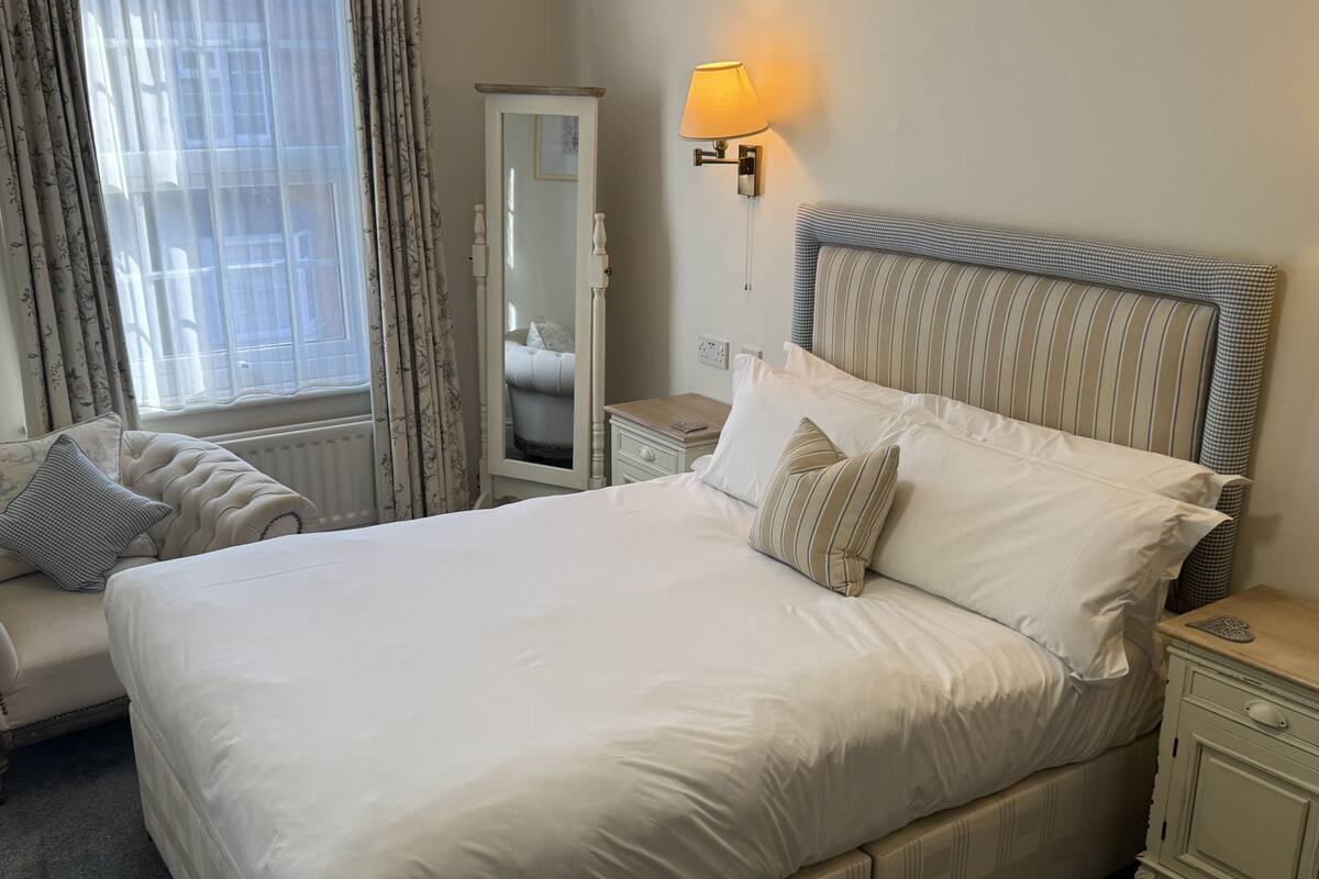 A hotel room with a large bed, a bedside table, a nightstand, a mirror, and a window with curtains.