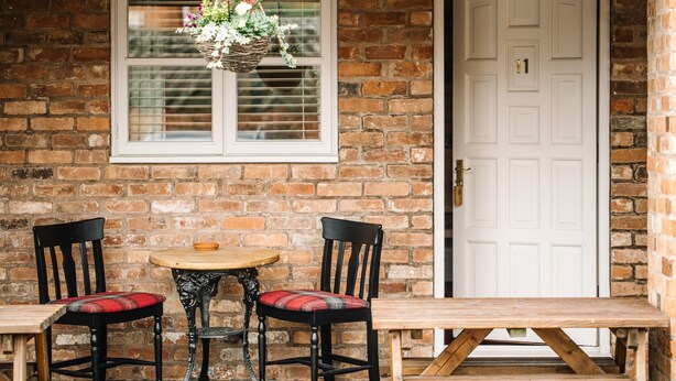 Een bakstenen muur, een witte deur met een messing knop, twee zwarte stoelen met kussens in rode en grijze strepen, een houten tafel en een hangmand met bloemen.