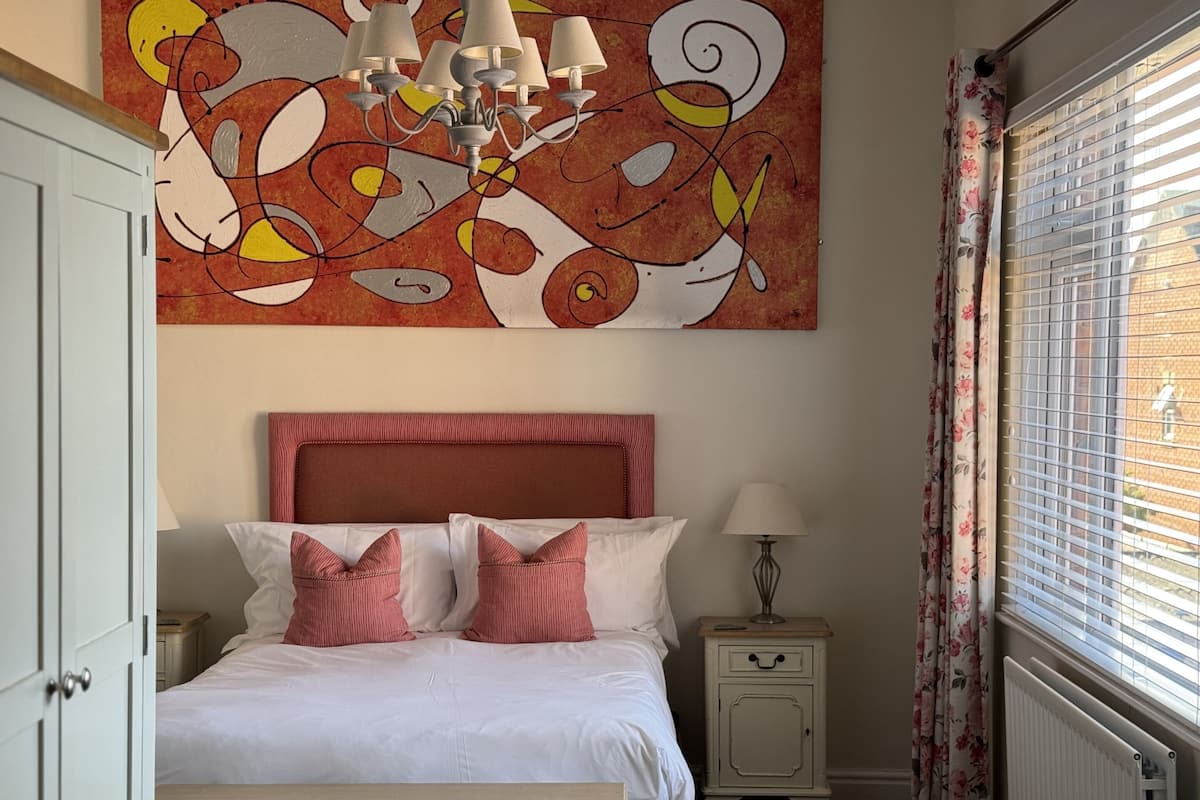 A bedroom with a bed, a nightstand, a lamp, a radiator, and a window with blinds.