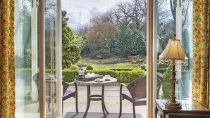 Un invernadero con una mesa de comedor y sillas, una lámpara y vistas a un jardín con un estanque y bancos.