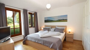 A bedroom with a bed, a TV, a bedside table, a lamp, and a window with curtains. - Beim Bicher (Grabenstätt)