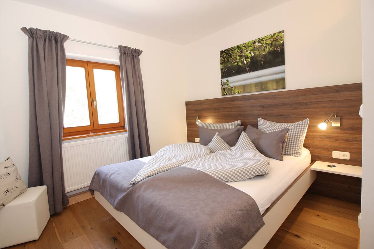 A bedroom with a bed, a window with curtains, a headboard, bedside lamps, and a wall-mounted artwork.