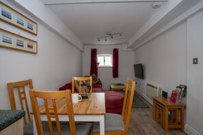 A cozy room with a wooden dining table, chairs, a red sofa, a small wooden side table, and a TV on a stand. - The Maltings (Canterbury)