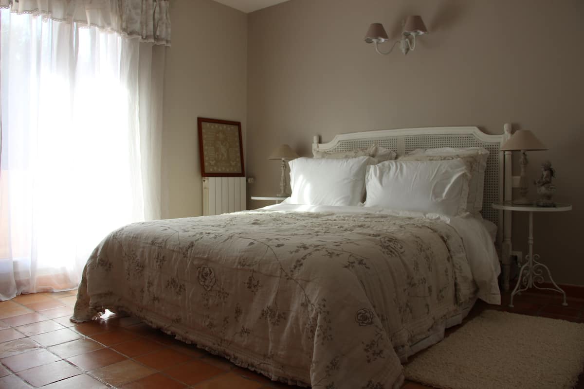 Une chambre à coucher avec un grand lit, une table de chevet avec une lampe et une fenêtre ornée de rideaux légers.