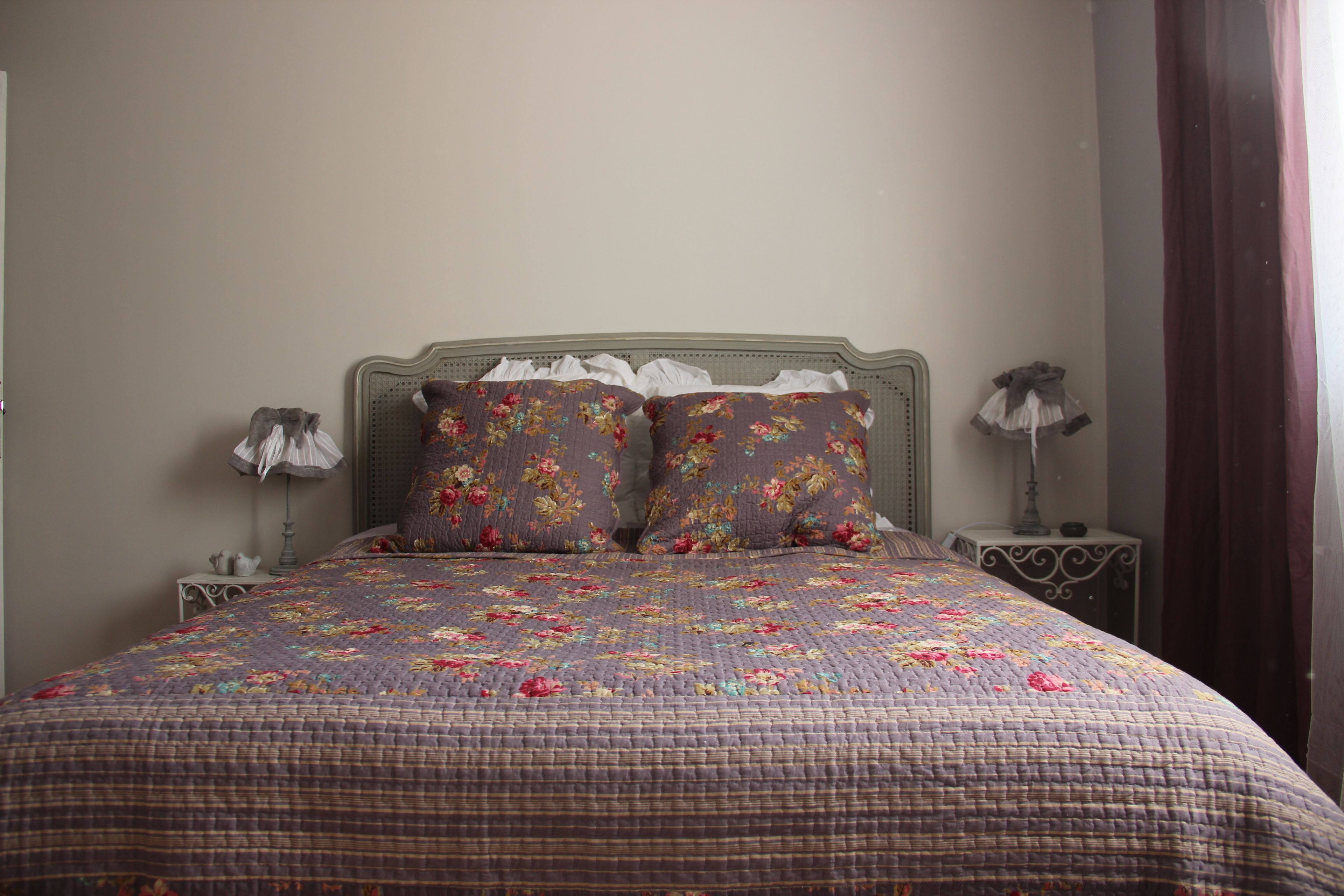 A bedroom with a floral-patterned bedspread, two matching pillows, a wooden headboard, two bedside tables with lamps, and a window with curtains.
