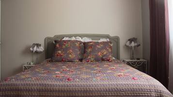 A bedroom with a floral-patterned bedspread, two matching pillows, a wooden headboard, two bedside tables with lamps, and a window with curtains.