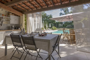 Un área de comedor al aire libre con una mesa y sillas, una piscina al fondo y un porche cubierto.