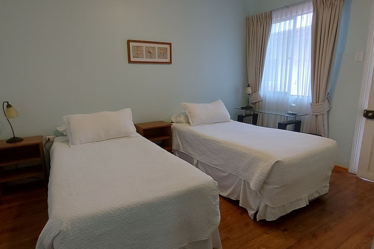 Una habitación con dos camas, una mesita de noche con lámpara, una pequeña mesa con lámpara, un cuadro en la pared y una ventana con cortinas.