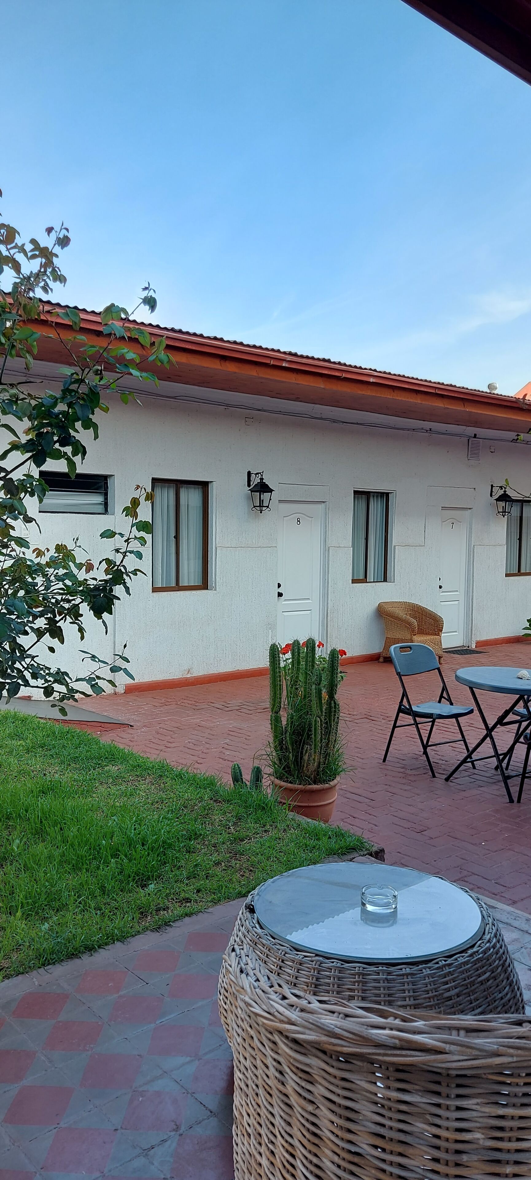 A building with a red-tiled roof, a white exterior, and a paved patio area with a wicker table and chairs, a potted cactus, and a small garden.