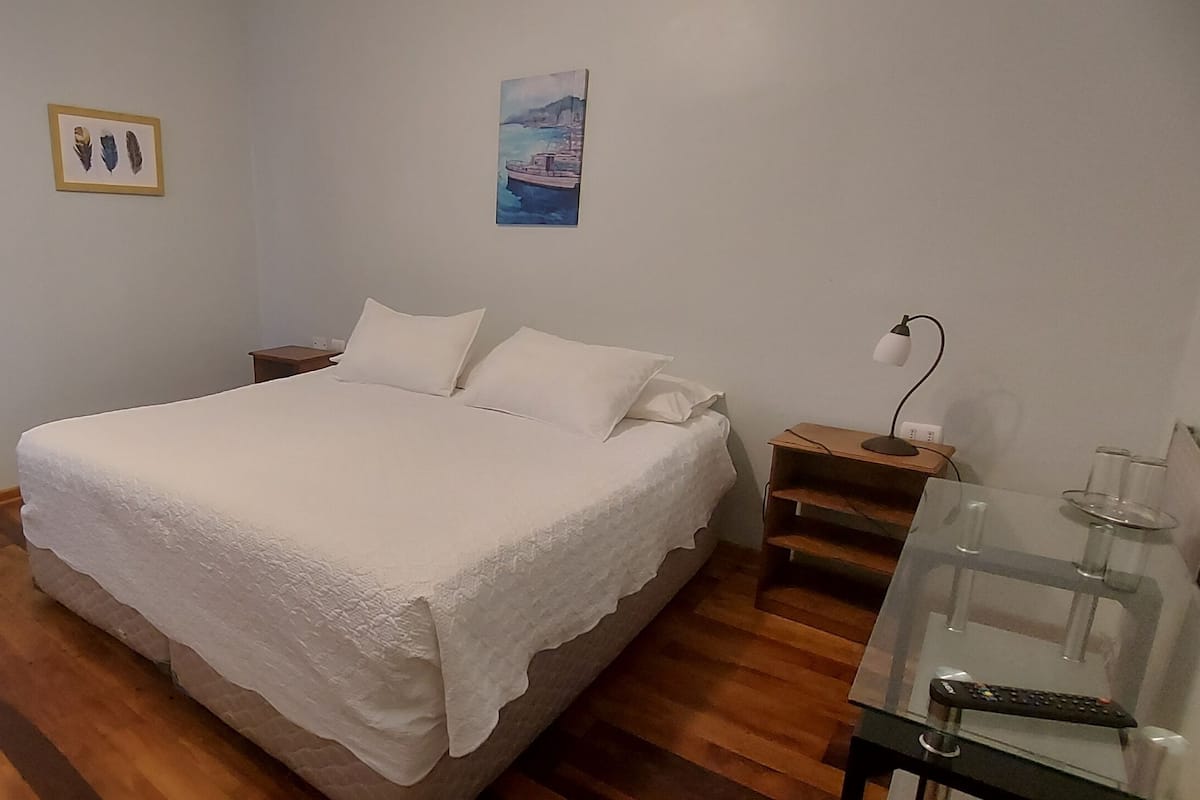 Un dormitorio con una cama grande, una mesita de noche, una silla y una mesa de vidrio con un control remoto.