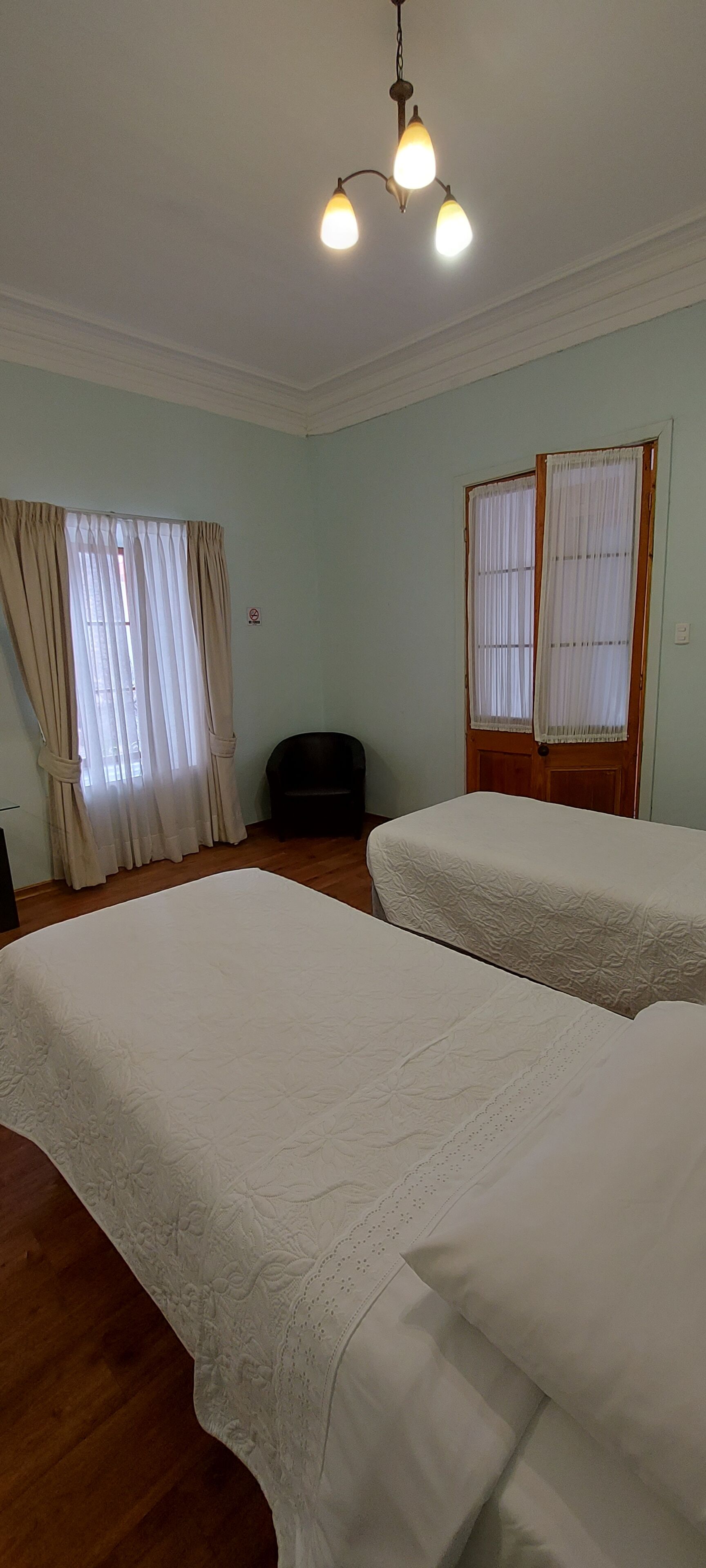 Quarto com duas camas, luminária no teto, janela com cortinas e uma porta.